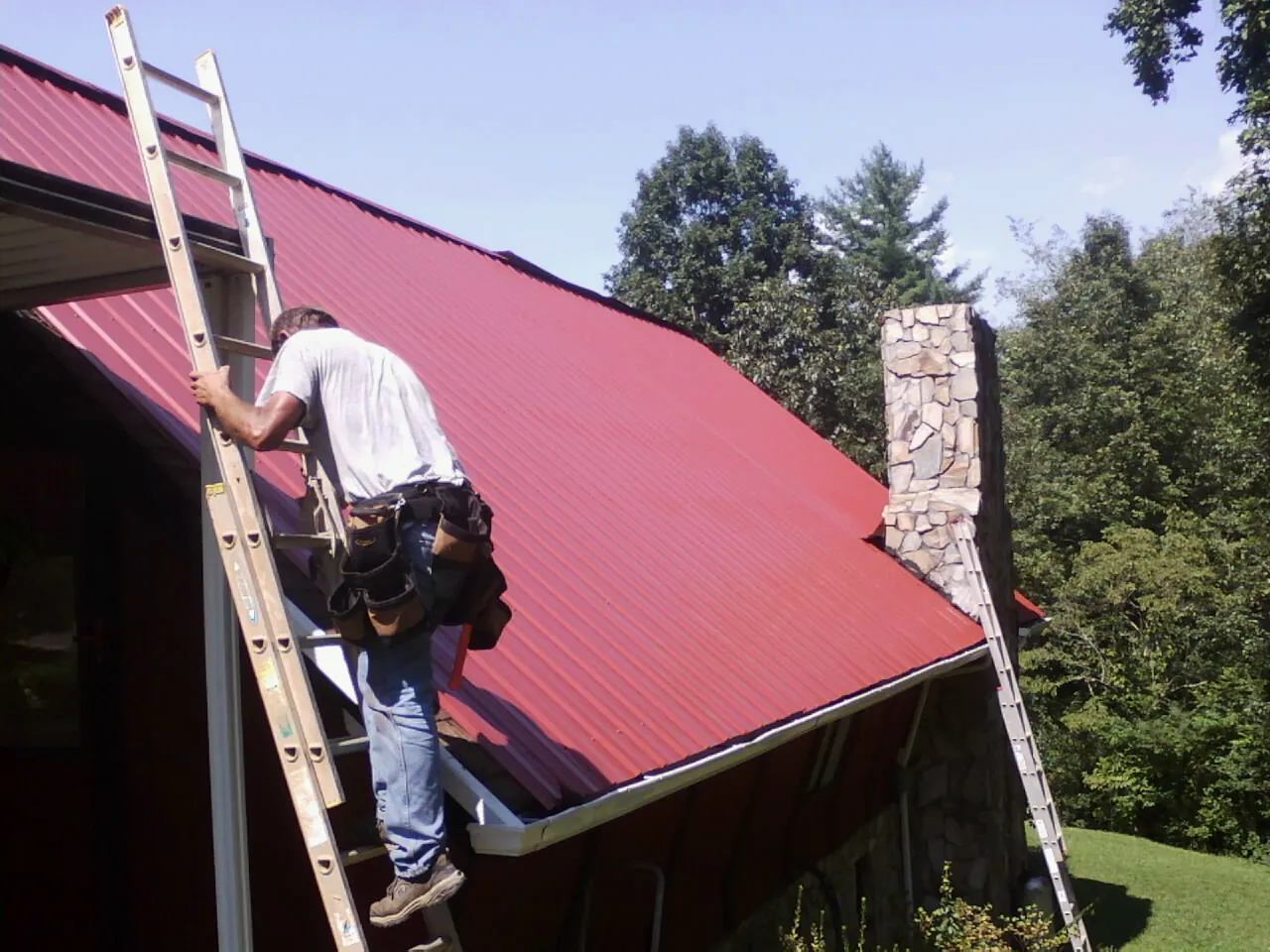 Roofer on a ladder beside a red metal roof and stone chimney — Soffit & Fascia Repair by Roofing Brothers in Neshanic Station